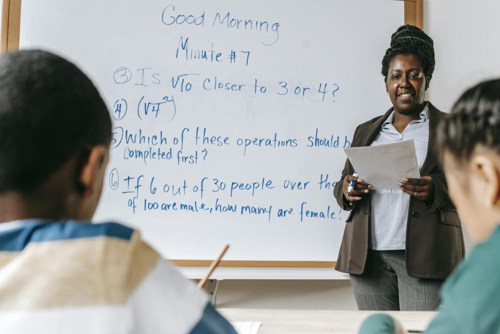 Cheerful African American female math teacher standing near whiteboard and explaining new material to multiracial pupils